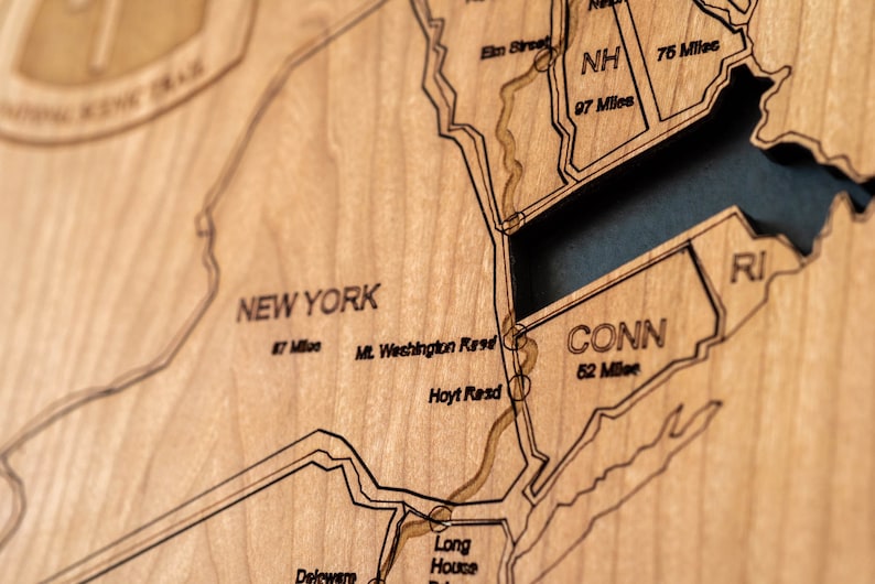 May include: Close-up of a wooden map of the northeastern United States, including New York, Connecticut, and Rhode Island. The map is etched with state lines, roads, and distances in miles. The wood has a light brown color.