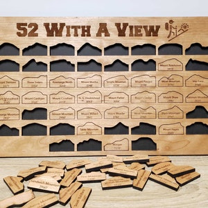 Könnte beinhalten: Ein Holzbrett mit dem Text "52 WITH A VIEW" und einem Wanderer-Symbol. Das Brett hat 52 Steckplätze für kleine Holzplättchen, jedes mit dem Namen und der Höhe eines Berges. Die Plättchen liegen verstreut auf einer Holzoberfläche.