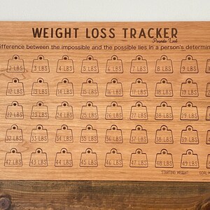 May include: A wooden weight loss tracker with 50 numbered squares, each representing a pound lost. The tracker is engraved with the quote "The difference between the impossible and the possible lies in a person's determination."  The words "Starting Weight" and "Goal Weight" are at the bottom of the tracker.