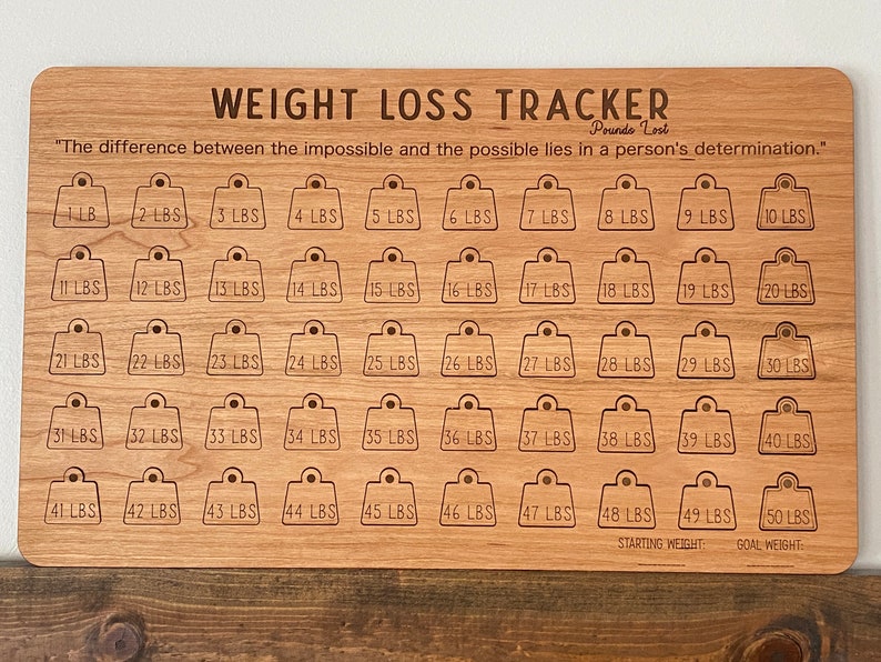 May include: A wooden weight loss tracker with 50 numbered squares, each representing a pound lost. The tracker is engraved with the quote "The difference between the impossible and the possible lies in a person's determination."  The words "Starting Weight" and "Goal Weight" are at the bottom of the tracker.