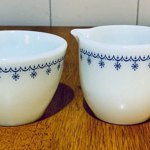 May include: A white ceramic creamer and sugar bowl set. The creamer has a handle and a spout. Both pieces have a blue snowflake and scallop design around the rim. The set is on a wooden surface.