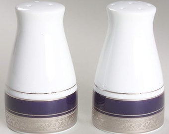 Op de afbeelding: Een paar witte keramische zout- en peperstrooiers. Elke strooier heeft een wit bovenstuk en een donkerblauwe band met een zilveren decoratieve rand. Een zilveren band scheidt de witte en blauwe secties.