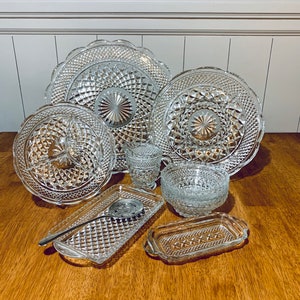 May include: A collection of clear, pressed glass serving dishes. The set includes plates, bowls, a serving tray with a spoon, and a butter dish. The glass has a diamond pattern and scalloped edges, creating a vintage aesthetic.