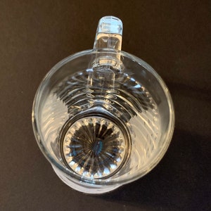 Peut inclure: Pichet en verre transparent avec une anse, vue de dessus. Le pichet a un corps large et rond avec un motif textur&eacute; et une base d&eacute;taill&eacute;e. La poign&eacute;e est rectangulaire et fix&eacute;e au bord sup&eacute;rieur. Le pichet est sur une surface sombre.