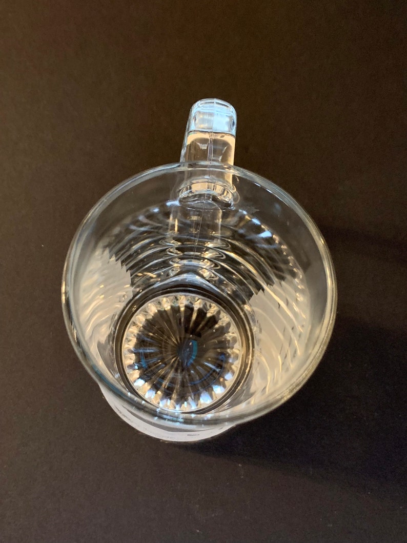 Peut inclure: Pichet en verre transparent avec une anse, vue de dessus. Le pichet a un corps large et rond avec un motif textur&eacute; et une base d&eacute;taill&eacute;e. La poign&eacute;e est rectangulaire et fix&eacute;e au bord sup&eacute;rieur. Le pichet est sur une surface sombre.