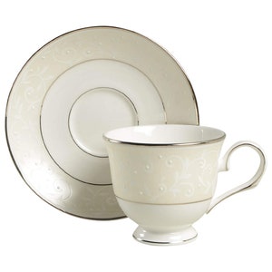 May include: Cream-colored teacup and saucer set with silver trim. The cup and saucer feature a delicate floral and dot pattern. The cup has a small handle and sits on a pedestal base. The saucer has a matching design.
