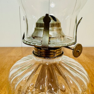 May include: Clear glass oil lamp with a ribbed base and a clear glass chimney. The lamp has a brass-colored metal top and a wick adjustment knob. The lamp is designed for use with oil and provides a classic lighting option.