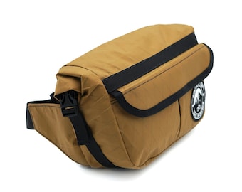 May include: A brown fanny pack with a black strap and buckle. The bag has a black zippered pocket on the front and a black patch with a white logo.
