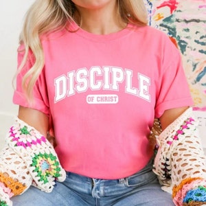 Op de afbeelding: Een roze T-shirt met witte tekst die "Disciple of Christ" zegt in een college-lettertype.