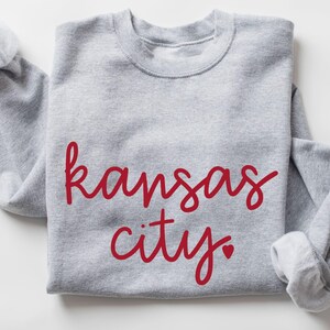 Puede incluir: Una sudadera gris con el texto "kansas city" en letras rojas con un pequeño corazón rojo.