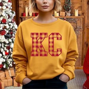 Op de afbeelding: Een mosterdgeel sweatshirt met de letters "KC" in rood en geel buffalo plaid.