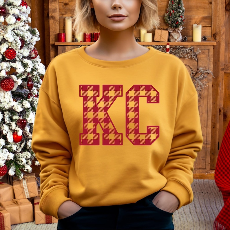 Op de afbeelding: Een mosterdgeel sweatshirt met de letters "KC" in rood en geel buffalo plaid.