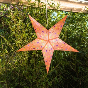 May include: An orange paper star lantern, decorated with small, colorful floral patterns. The star is suspended by a white string against a backdrop of green bamboo and wooden beams. A decorative item for the home.