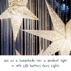 May include: Two white paper star lanterns with intricate cut-out designs. The lanterns are illuminated and hanging from a string of white beads. The text "use as a lampshade over a pendant light or with LED battery fairy lights" is written below the lanterns.