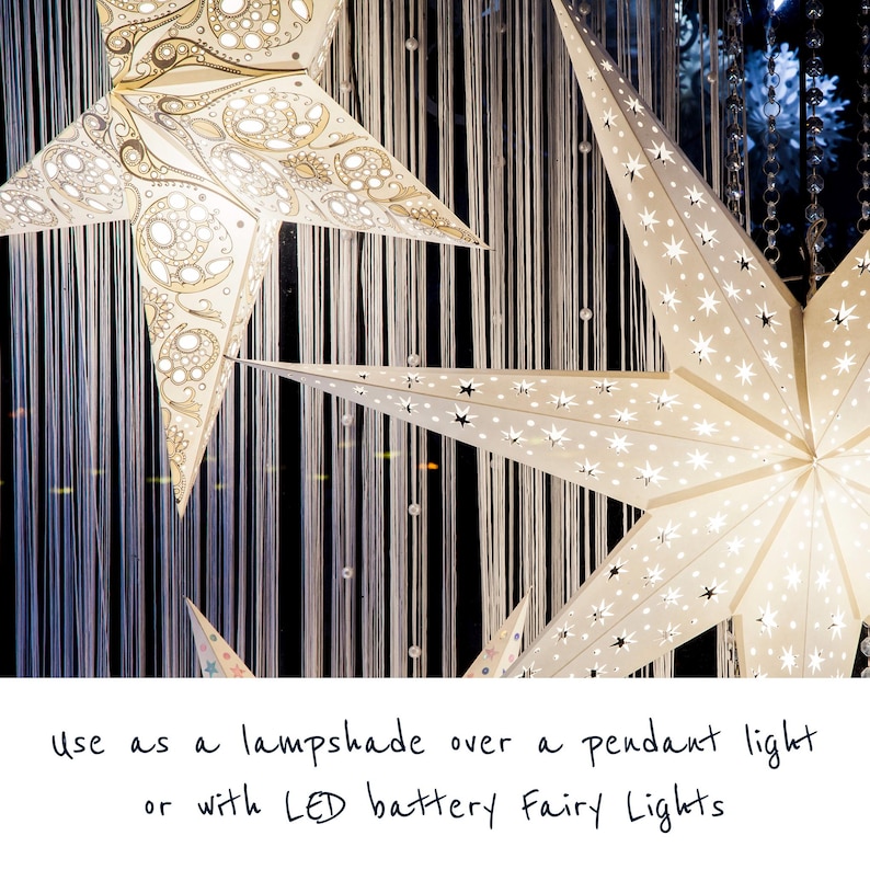 May include: Two white paper star lanterns with intricate cut-out designs. The lanterns are illuminated and hanging from a string of white beads. The text "use as a lampshade over a pendant light or with LED battery fairy lights" is written below the lanterns.