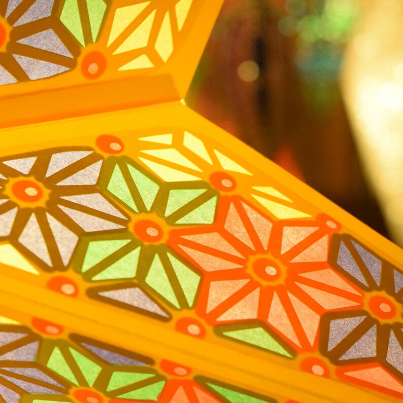 K&ouml;nnte beinhalten: Nahaufnahme einer gelben, sternf&ouml;rmigen Papierlaterne mit einem geometrischen Muster aus blauen, gr&uuml;nen, orangefarbenen und rosa Bl&uuml;ten. Die Laterne wird von hinten beleuchtet, wodurch ein leuchtender Effekt entsteht.
