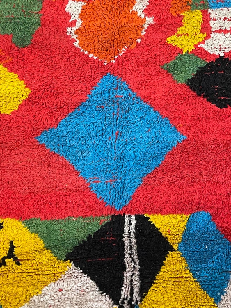 May include: A colorful rug with geometric shapes in shades of red, blue, yellow, green, black, and white. The rug has a fuzzy texture.