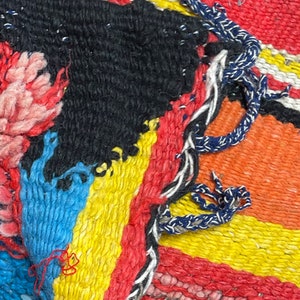May include: Close-up of a colorful woven rug with a variety of textures and patterns. The rug is made of different colored yarns, including red, orange, yellow, blue, and black. The rug has a frayed edge and a knotted fringe.