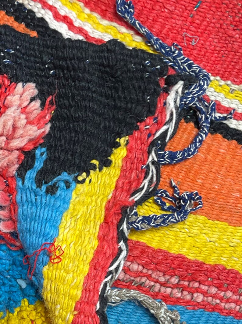 May include: Close-up of a colorful woven rug with a variety of textures and patterns. The rug is made of different colored yarns, including red, orange, yellow, blue, and black. The rug has a frayed edge and a knotted fringe.