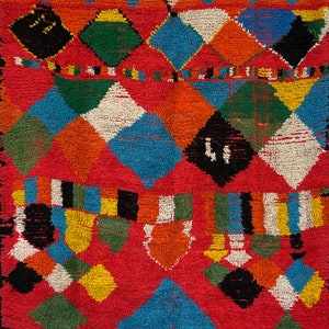 May include: A colorful Moroccan rug with a geometric pattern in shades of red, orange, blue, green, white, and black. The rug is made of wool and has a soft, fuzzy texture.