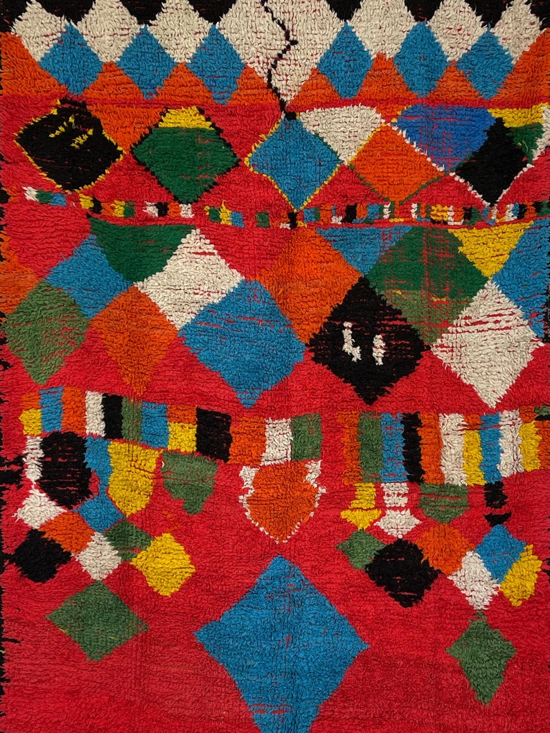 May include: A colorful Moroccan rug with a geometric pattern in shades of red, orange, blue, green, white, and black. The rug is made of wool and has a soft, fuzzy texture.