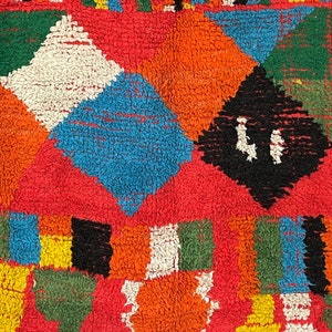 May include: A colorful rug with a geometric pattern in shades of red, orange, blue, green, white, and black. The rug is made of a soft, fluffy material.