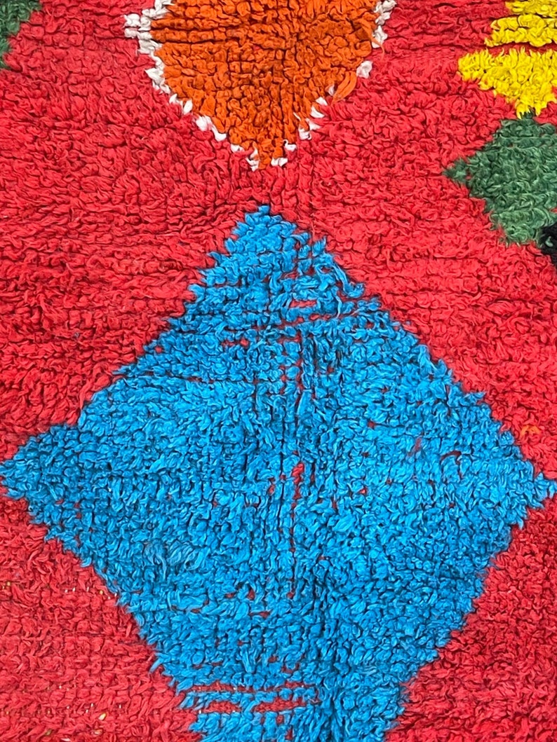 May include: A close-up of a colorful rug with a diamond pattern. The rug is made of a soft, fuzzy material and features shades of red, blue, orange, yellow, and green.