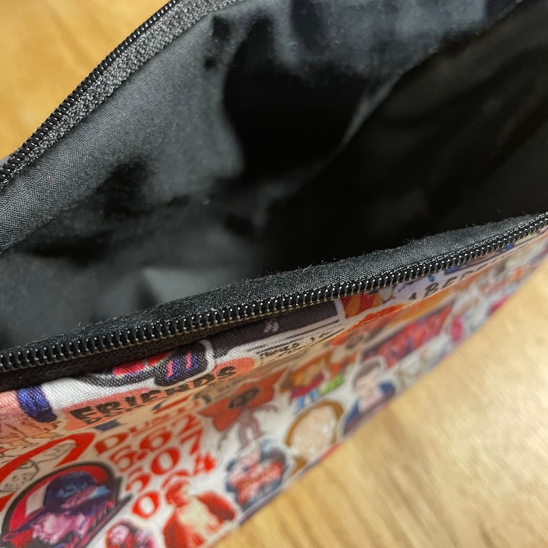 Puede incluir: Bolsa con cremallera abierta con interior negro y exterior blanco cubierto de im&aacute;genes y texto coloridos. Las im&aacute;genes incluyen personajes y logotipos de la serie de televisi&oacute;n Friends.