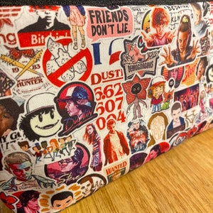 Puede incluir: Una bolsa de tela blanca cubierta de coloridas pegatinas con personajes y frases de la serie de televisi&oacute;n "Stranger Things". Las pegatinas incluyen im&aacute;genes de personajes, el Demogorgon y el texto "FRIENDS DON'T LIE."