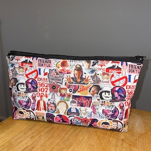 Puede incluir: Un neceser blanco con cremallera, cubierto de coloridas pegatinas con personajes y frases de la serie de televisi&oacute;n Stranger Things. El neceser tiene una cremallera negra y est&aacute; sobre una superficie de madera.