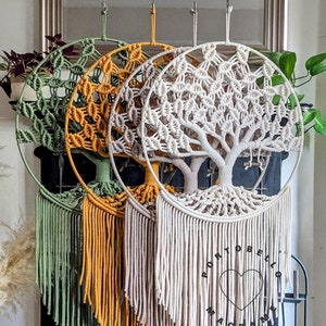 May include: Three macrame wall hangings in the shape of a tree of life. The hangings are in green, yellow, and white. The hangings are hanging in front of a fireplace. The bottom of the hangings have long fringe. The white hanging has a black circle with a heart and the text "Portobello Macrame Art" in the center.