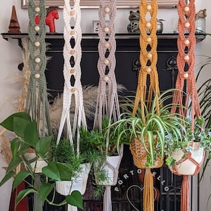 May include: Five macrame plant hangers with wooden beads and tassels in green, white, grey, yellow, and orange hanging from a fireplace mantel.