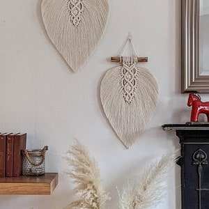 Puede incluir: Dos tapices de macram&eacute; beige en forma de hojas con listones de madera en la parte superior. Los tapices est&aacute;n en una pared blanca con un estante de madera debajo.