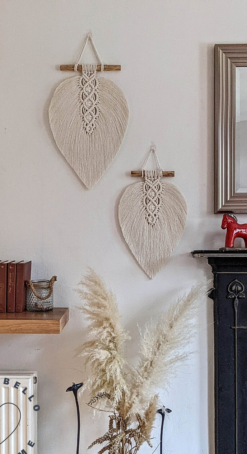 Puede incluir: Dos tapices de macram&eacute; beige en forma de hojas con listones de madera en la parte superior. Los tapices est&aacute;n en una pared blanca con un estante de madera debajo.