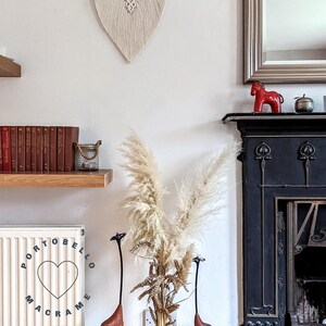Puede incluir: Un tapiz de macram&eacute; blanco en forma de hoja con una barra de madera en la parte superior. El tapiz est&aacute; colgado en una pared blanca. Hay una chimenea negra con una figurita de caballo de cer&aacute;mica roja en la repisa. Hay un jarr&oacute;n con hierba de la pampa seca y dos esculturas de jirafas altas en primer plano.