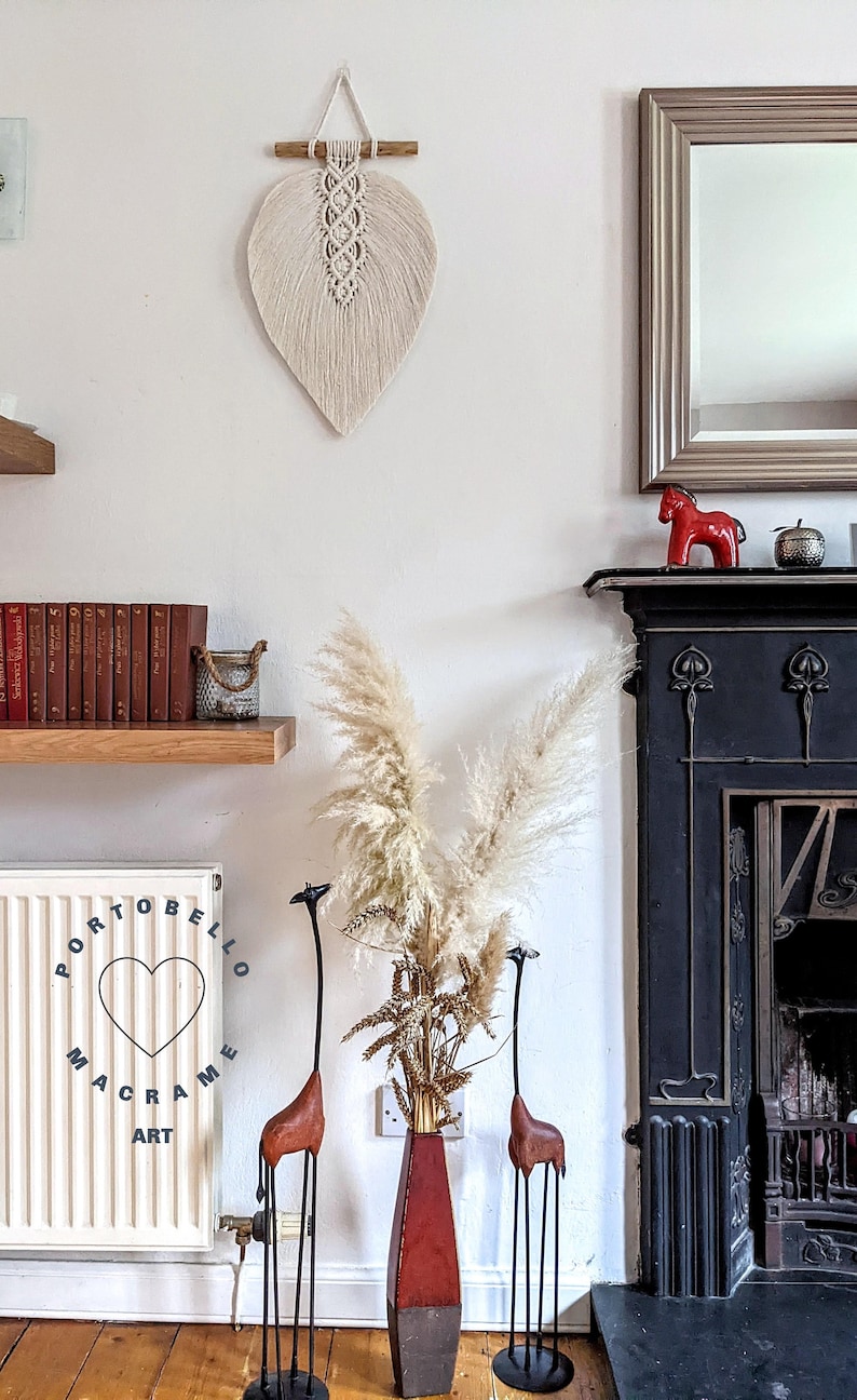 Puede incluir: Un tapiz de macram&eacute; blanco en forma de hoja con una barra de madera en la parte superior. El tapiz est&aacute; colgado en una pared blanca. Hay una chimenea negra con una figurita de caballo de cer&aacute;mica roja en la repisa. Hay un jarr&oacute;n con hierba de la pampa seca y dos esculturas de jirafas altas en primer plano.
