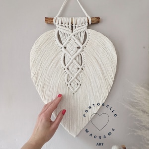 May include: White macrame wall hanging in the shape of a heart with a wooden dowel at the top. The macrame is made with a knot pattern and has a fringe at the bottom. The text "Portobello Macrame Art" is printed below the hanging.