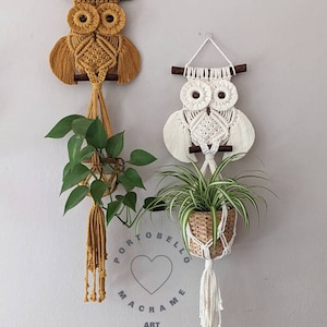 Könnte beinhalten: Zwei Makramee-Wandbehänge in Form von Eulen, einer in Gelb und einer in Weiß, mit einer Pflanze, die an jedem hängt. Der Text "Portobello Macrame Art" ist unter den Eulen sichtbar.