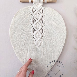 Puede incluir: Un colgante de pared de macram&eacute; en forma de hoja blanca con una barra de madera en la parte superior. La hoja est&aacute; hecha con una t&eacute;cnica de anudado y tiene una franja en la parte inferior. El colgante tiene un logotipo que dice "Portobello Macrame Art" con un coraz&oacute;n en el centro.