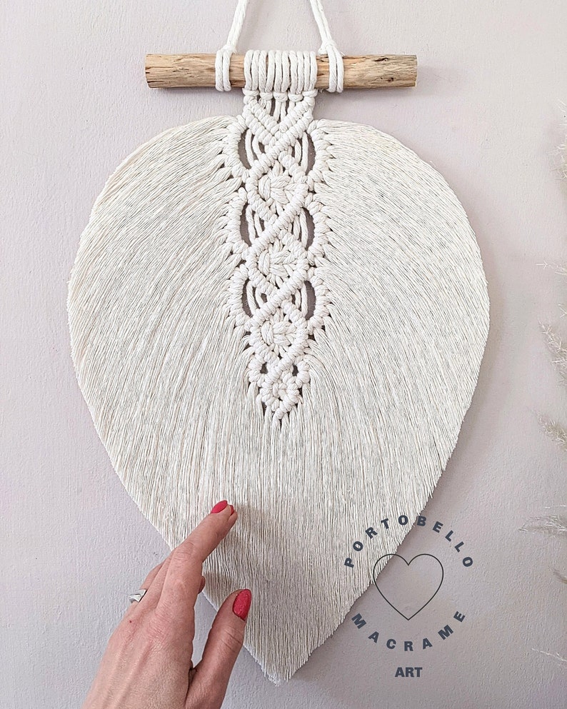 Puede incluir: Un colgante de pared de macram&eacute; en forma de hoja blanca con una barra de madera en la parte superior. La hoja est&aacute; hecha con una t&eacute;cnica de anudado y tiene una franja en la parte inferior. El colgante tiene un logotipo que dice "Portobello Macrame Art" con un coraz&oacute;n en el centro.
