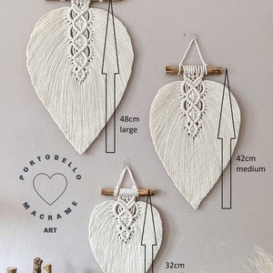 Puede incluir: Tres colgantes de pared de macram&eacute; en forma de hoja de color blanco, de diferentes tama&ntilde;os. El m&aacute;s grande mide 48 cm de alto, el mediano 42 cm y el m&aacute;s peque&ntilde;o 32 cm. El texto "Portobello Macrame Art" es visible en la esquina inferior izquierda de la imagen.