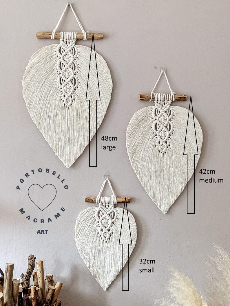 Puede incluir: Tres colgantes de pared de macram&eacute; en forma de hoja de color blanco, de diferentes tama&ntilde;os. El m&aacute;s grande mide 48 cm de alto, el mediano 42 cm y el m&aacute;s peque&ntilde;o 32 cm. El texto "Portobello Macrame Art" es visible en la esquina inferior izquierda de la imagen.