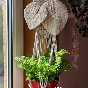 Peut inclure: Un porte-plante en macramé blanc avec deux motifs en forme de plumes est suspendu à une baguette en bois. Une plante verte dans un pot rouge est suspendue au porte-plante. Le porte-plante a un pompon blanc en bas et le texte "Portobello Macrame Art" est imprimé sur le pot.