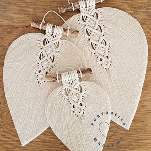 Puede incluir: Tres tapices de macram&eacute; con forma de hoja de diferentes tama&ntilde;os, cada uno con una varilla de madera natural en la parte superior. La hoja m&aacute;s grande tiene el texto "Portobello Macrame Art" impreso en ella.