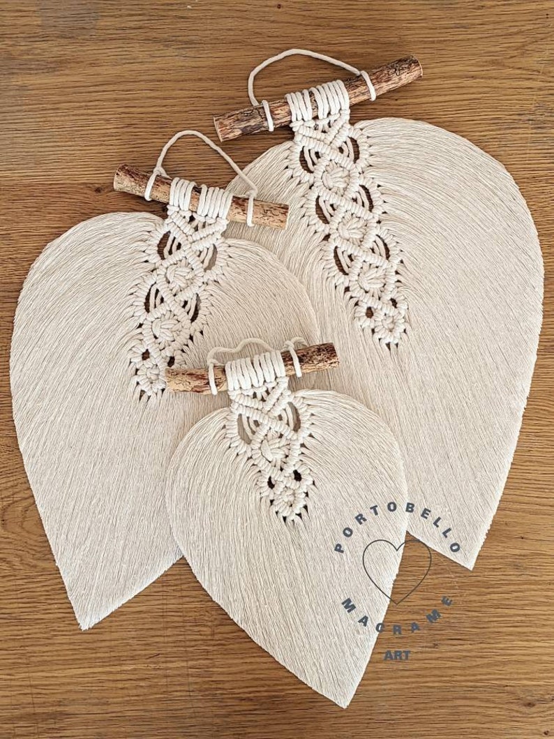 Puede incluir: Tres tapices de macram&eacute; con forma de hoja de diferentes tama&ntilde;os, cada uno con una varilla de madera natural en la parte superior. La hoja m&aacute;s grande tiene el texto "Portobello Macrame Art" impreso en ella.