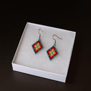 Peut inclure: Une paire de boucles d'oreilles losanges perlées. Les boucles d'oreilles présentent un motif rouge, jaune et orange au centre, entouré de perles turquoise. Elles sont présentées dans une boîte blanche sur fond noir.