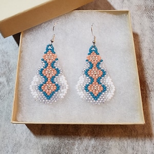 May include: A pair of teardrop-shaped earrings with a geometric pattern of white, peach, and teal beads. The earrings are hanging from silver hooks.