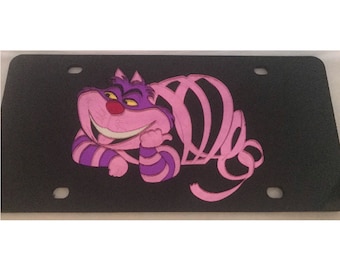 Op de afbeelding: Een zwart kenteken met een roze en paarse Cheshire Cat uit Alice in Wonderland. De kat grijnst en heeft een gestreept patroon.