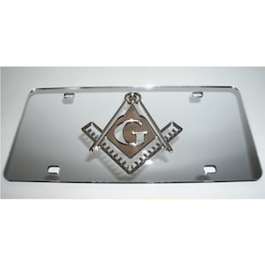 May include: A silver chrome license plate frame with a raised, silver Masonic symbol, including a square and compass with a 'G' in the center.