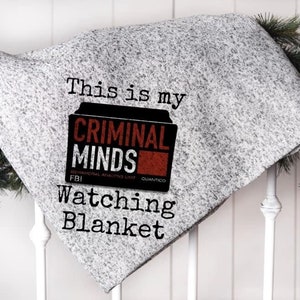 May include: A gray blanket with the text "This is my CRIMINAL MINDS Watching Blanket." The blanket has a graphic of a file folder with the words "CRIMINAL MINDS" and "FBI" on it. The blanket is draped over a white railing.